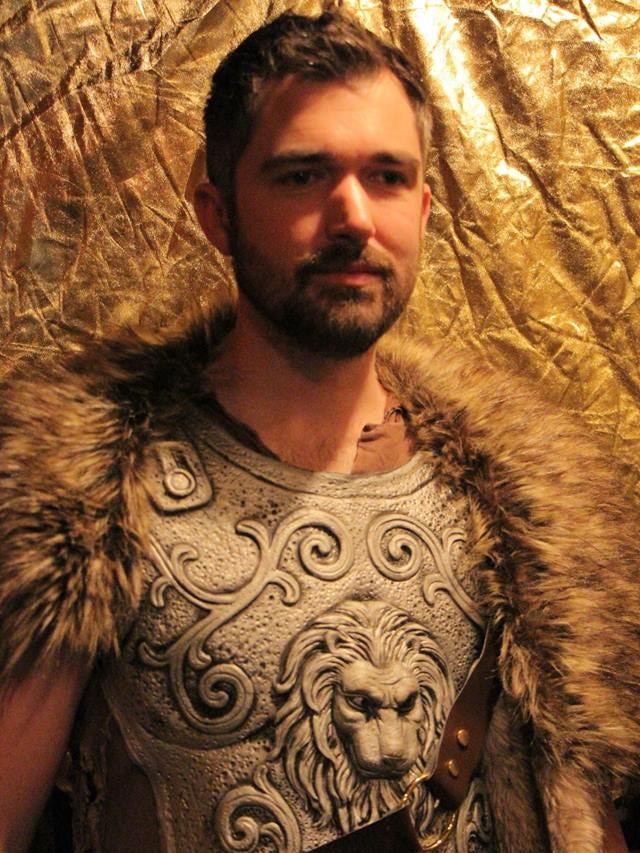 A man wearing a fancy dress costume of armour and a fur collar in front of some golden fabric.