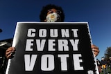 A woman wearing a mask holds a sign saying 'count every vote'.