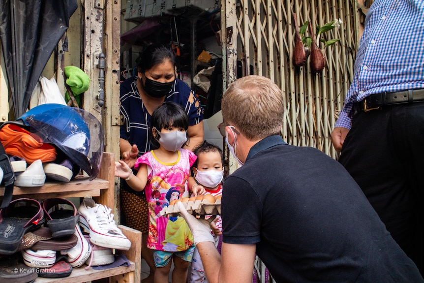 Two little Thai girls in a doorway look at a packet of eggs being held by a crouching man. 