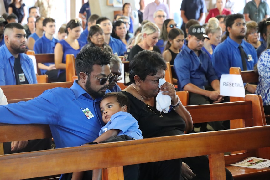A man holds a small child and sits next to a woman wiping her eyes with a tissue. They are sitting on church pews.