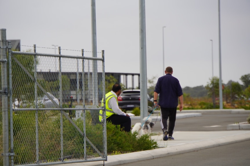 A security guard sits at a gate as a man walks his dogs at the entrance to the Bullsbrook COVID quarantine centre.