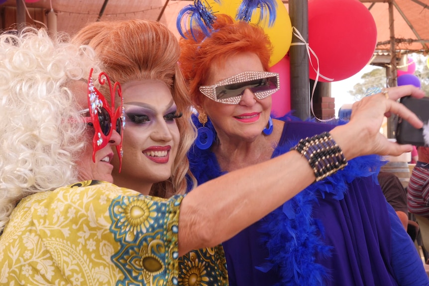 A drag queen poses for a selfie with two colourfully dressed women.