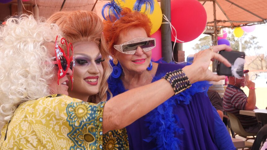 A drag queen poses for a selfie with two colourfully dressed women.
