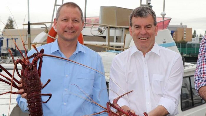 Two men in white and blue shirts hold up rock lobsters in front of a boat.