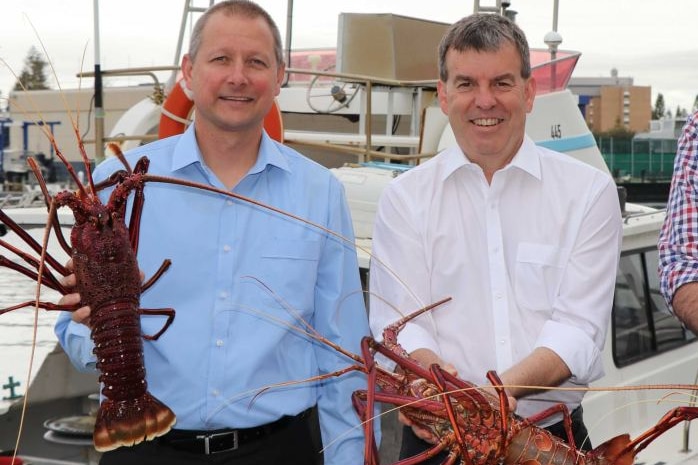 Two men in white and blue shirts hold up rock lobsters in front of a boat.