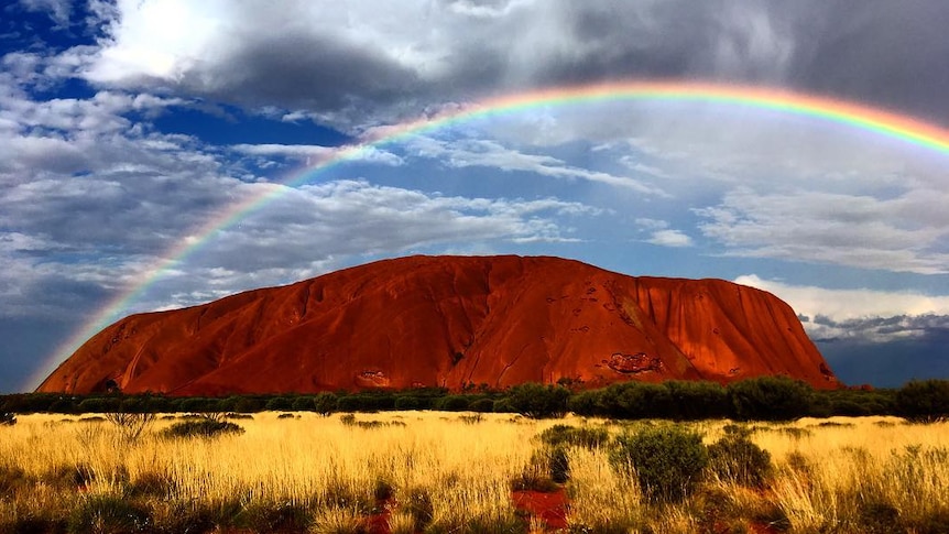Large dark orange rock with rainbow arching over the top and cloudy sky