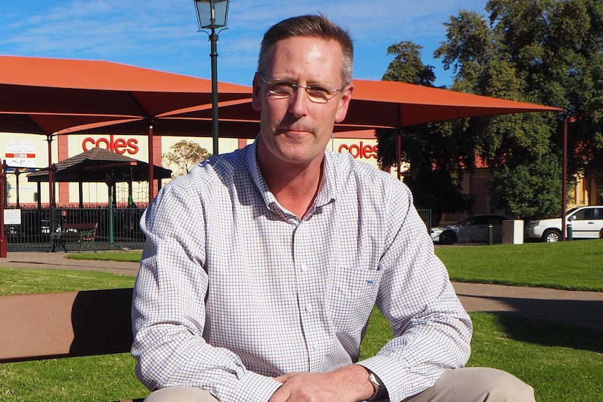 A tall man in a checked white shirt sits on a park bench in a park covered in green grass with a playground in the background.