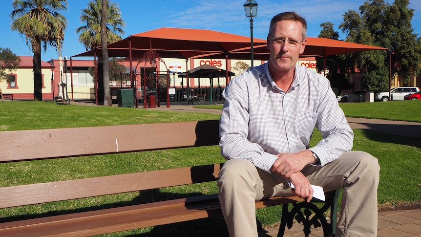 A tall man in a checked white shirt sits on a park bench in a park covered in green grass with a playground in the background