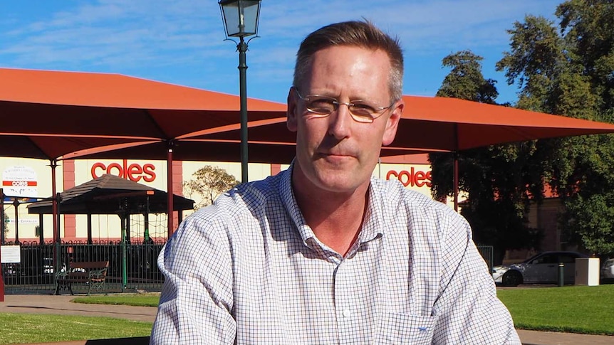 A tall man in a checked white shirt sits on a park bench in a park covered in green grass with a playground in the background