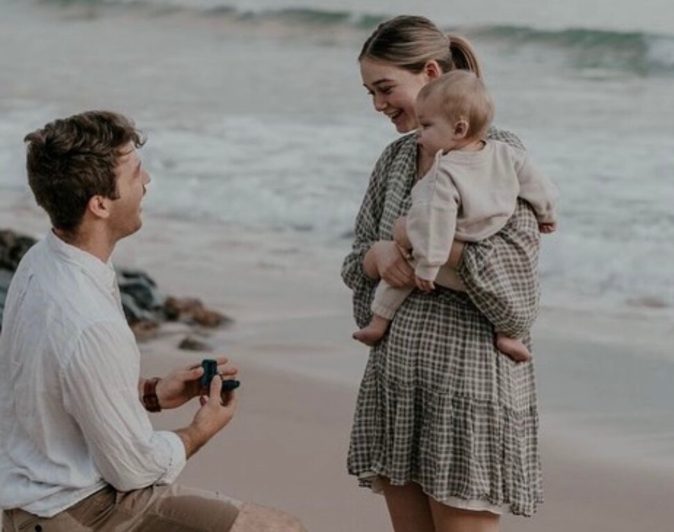 A man kneeling proposing to a woman holding a baby on the beach. They both smile as waves crash behind them.