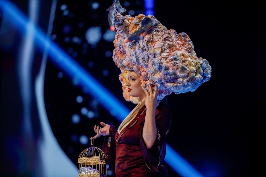 Jane Garrett wearing a headdress that looks like smoke and missiles, holding a fake dove in a cage