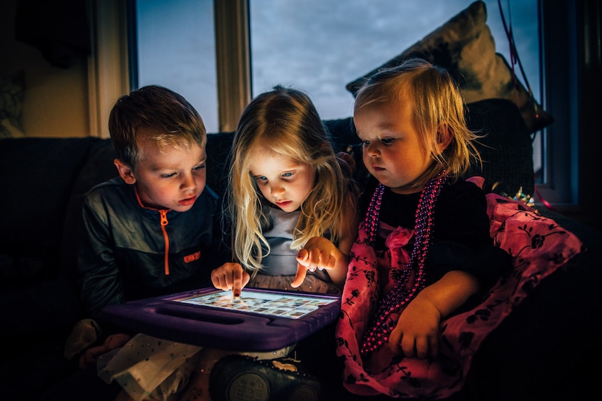 Three children on couch looking at tablet in story about kids and routines during coronavirus isolation.