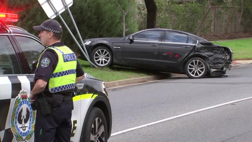 A damaged black Commodore halfway on a verge. A police officer stands next to a police car in the foreground