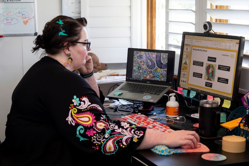 A woman wearing a black top with colourful embroidering along the arm sits at a desk with two screens.