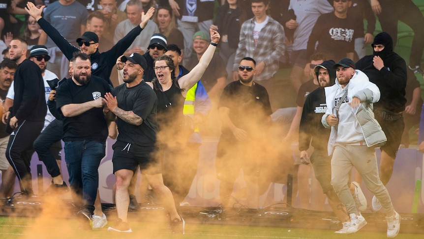 A group of people run onto a pitch, with orange haze from flares around them.
