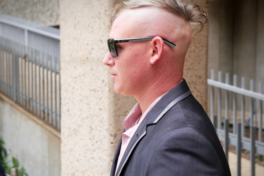A man  with a half-shaved head wearing sunglasses outside a courthouse 
