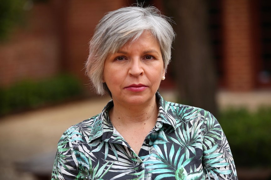 A woman in a blouse stands looking seriously at the camera.