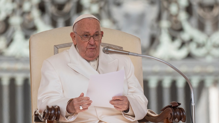 pope francis dressed in white suit and pope's hat sits in chair with microphone, holding paper in his hands