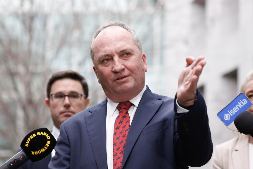 A man with receding grey hair, wearing a blue suit, red and black tie answering media questions with party members behind him