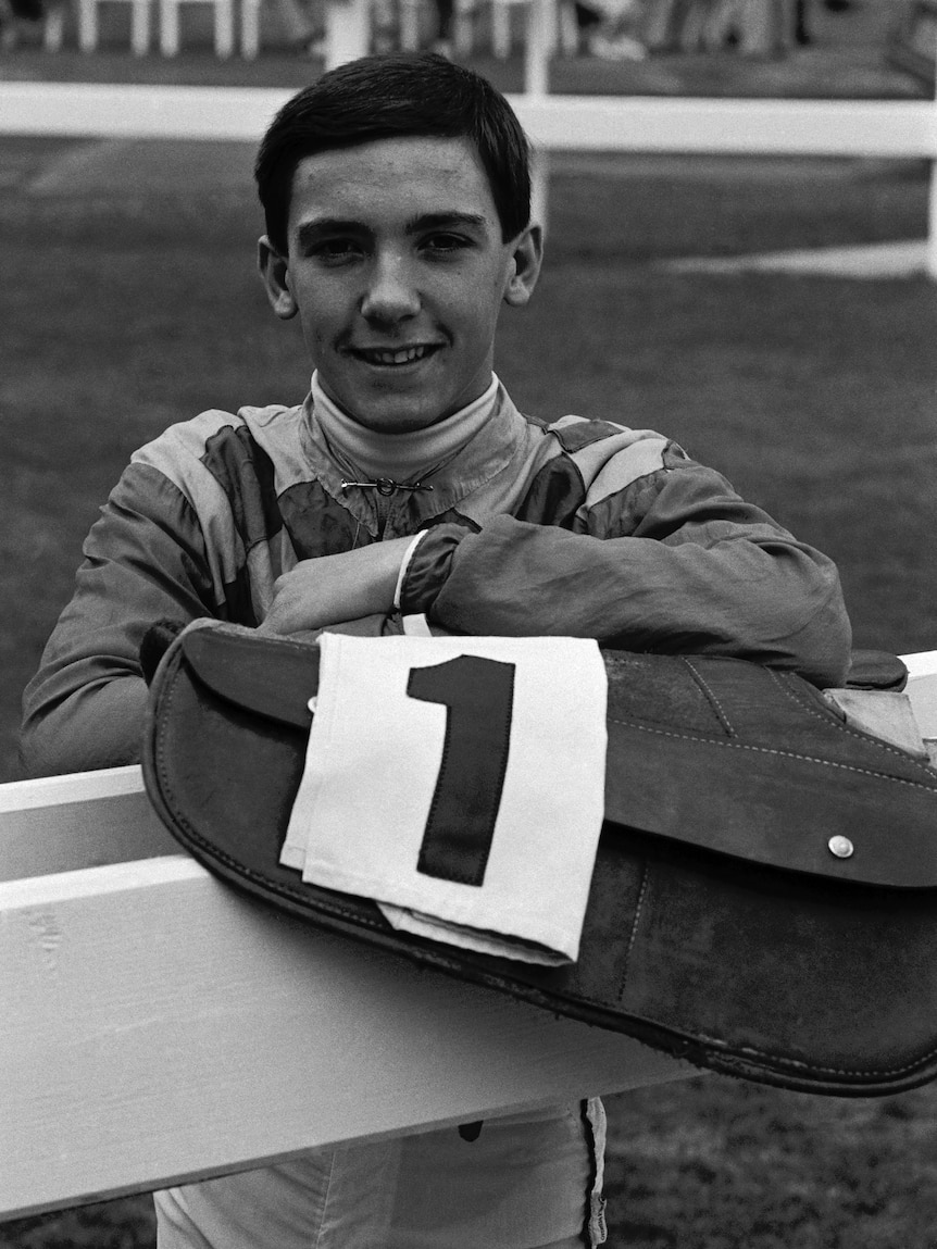 A young jockey stands in the mounting yard looking at the camera while holding his saddle.