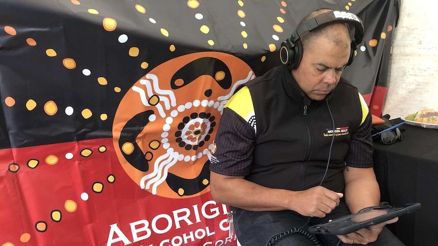 Aboriginal health worker sitting in front of ADAC sign using the Grog App with headphones on.