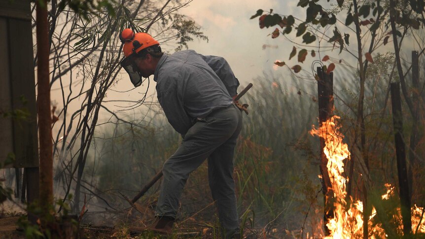 A mean wearing a firefighting helmet uses a rake to shift soil towards a fire burning in a grassy area.