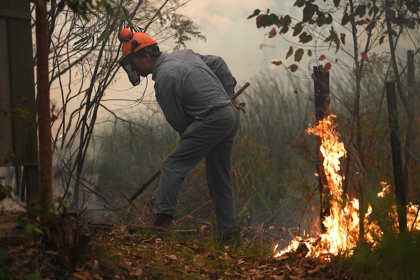 A mean wearing a firefighting helmet uses a rake to shift soil towards a fire burning in a grassy area.