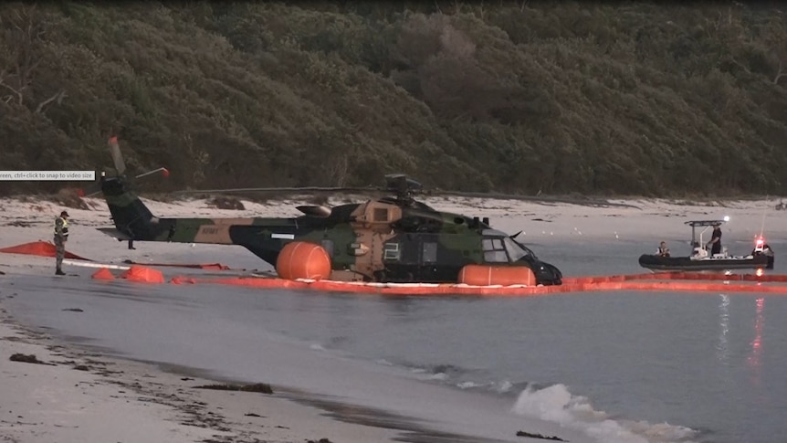 An image of an army helicopter in water near a beach as a small police boat approaches on the right side.