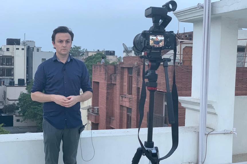 James Oaten in a navy shirt standing on a roof top looking into a camera on a tripod