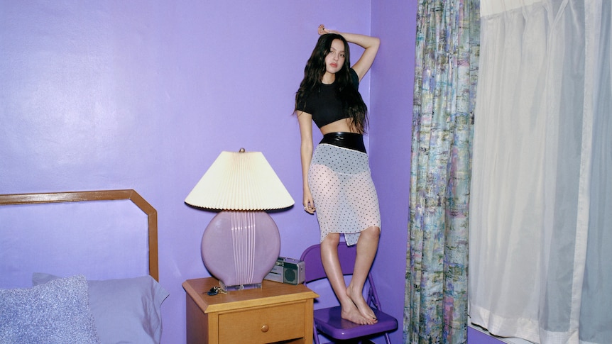 Olivia Rodrigo wears a black top and sheer white skirt and stands on a purple chair and leans against a purple bedroom wall