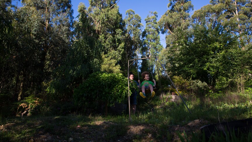A young mother pushes a little boy on a lop-sided, old-fashioned swing set surrounded by bush.