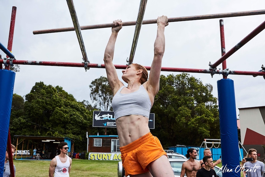 A muscular woman in white top and orange shorts grips onto a long stick as she navigates an obstacle course