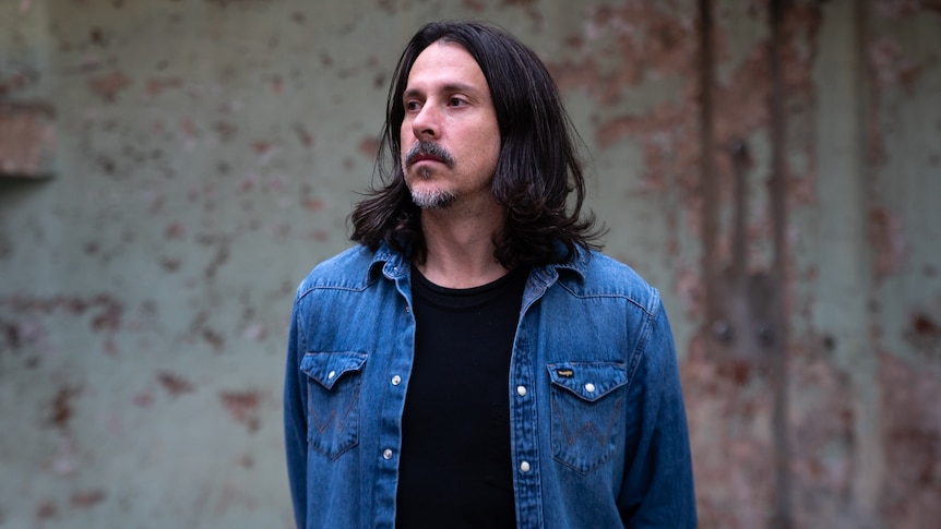 Man in denim jacket with shoulder-length brown hair and short beard looks serious 