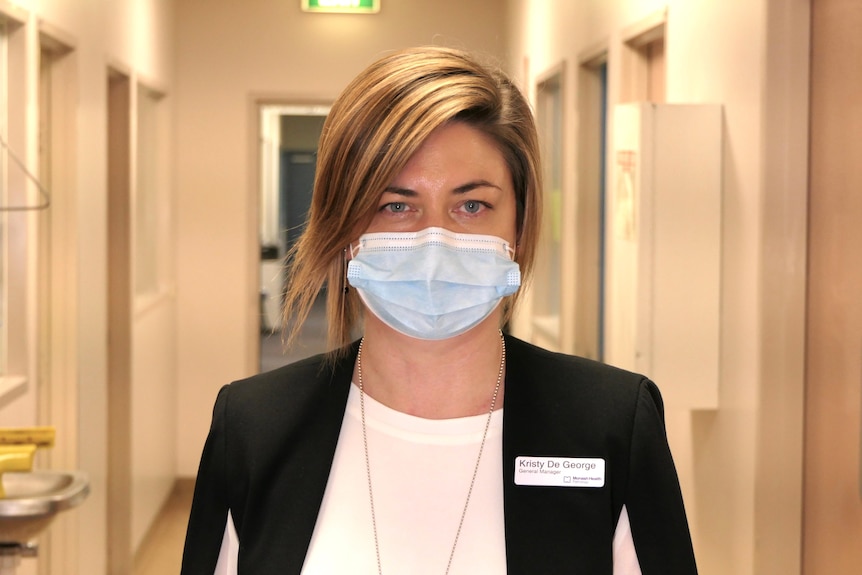 A fair-haired woman in a surgical mask looking at the camera. She is wearing a split-sleeve black jacket over a white top
