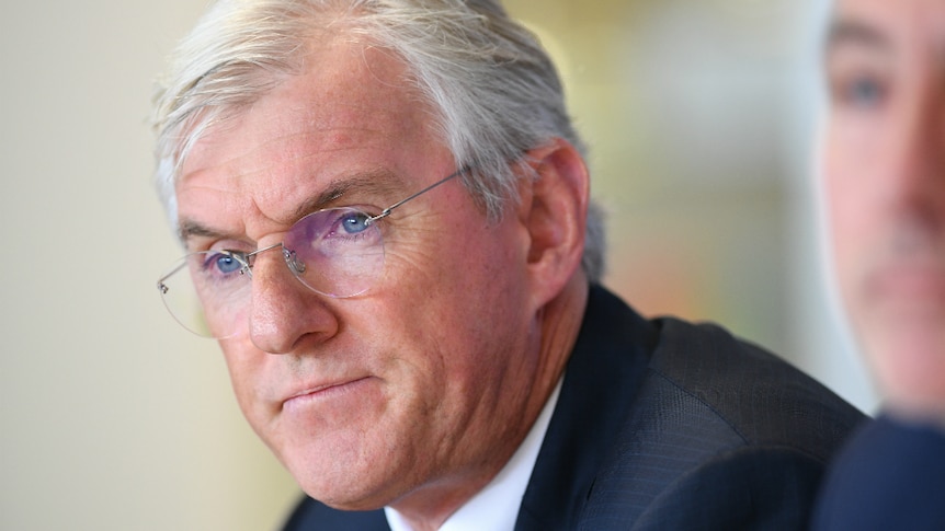 A silver haired white man in a suit and spectacles wears a neutral expression