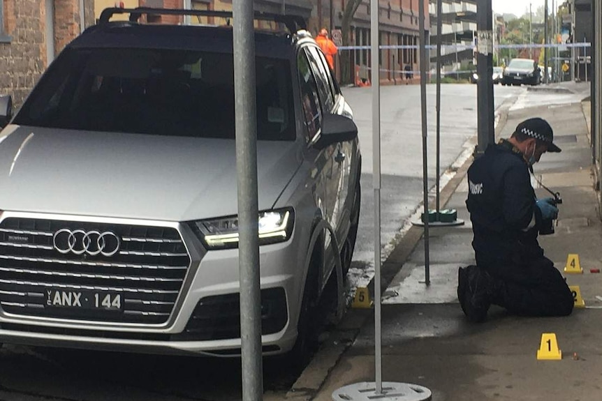 A police officer kneels on the ground taking photos and an Audi SUV is parked on the street under an SES marquee tent.