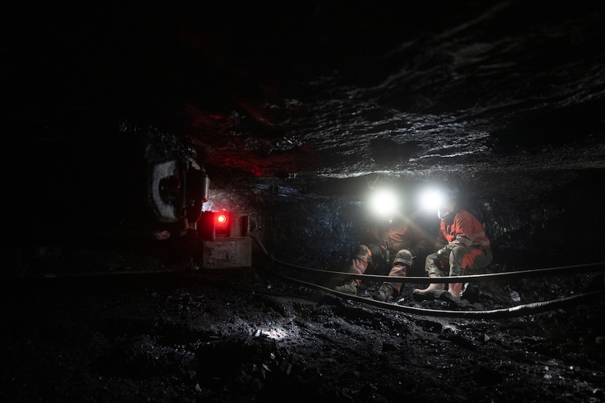 In the dark depths of a coal mine with low ceilings, two workers in headlamps are sitting together.