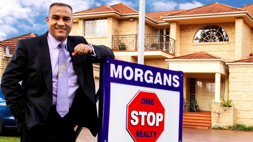 A man in a suit leans against a real estate sign in front of a house.