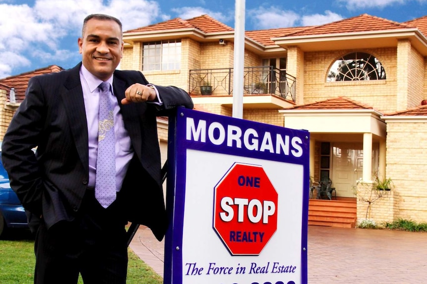 A man in a suit leans against a real estate sign in front of a house.