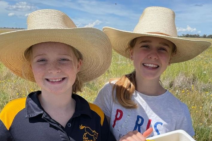 Two girls in akubras hold a tub of picked yellow wildflowers