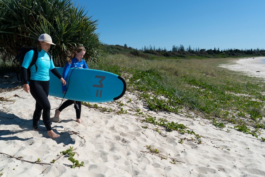 A woman and a child walk onto a beach with a surfboard for a story about surf therapy helping autistic children