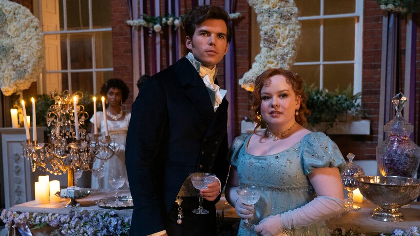 A man and a woman wearing old fashioned regency garments look toward the camera. She smiles and he looks nervous.
