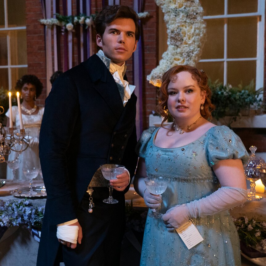 A man and a woman wearing old fashioned regency garments look toward the camera. She smiles and he looks nervous.