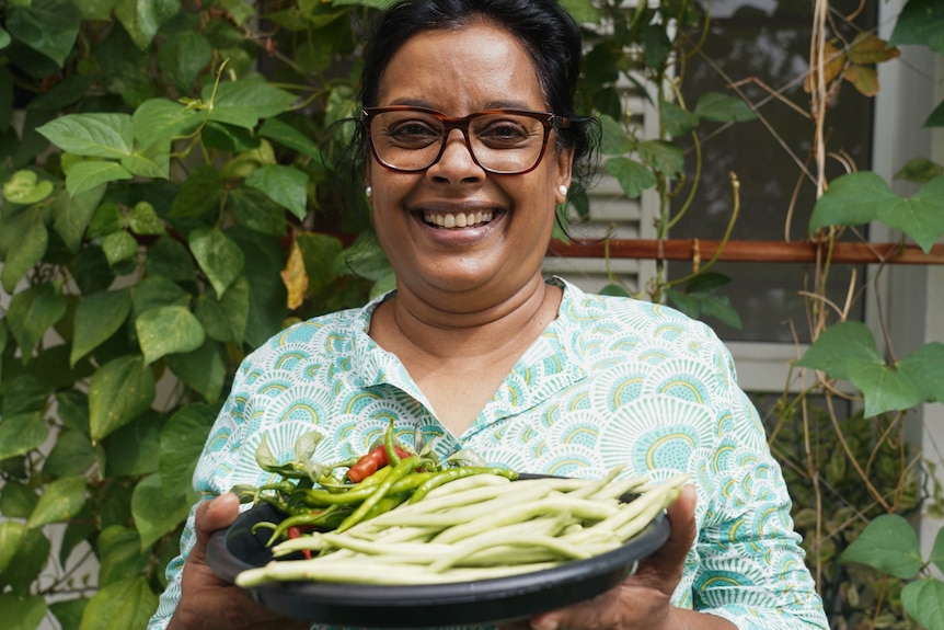 A woman in a green patterned shirt grins widely as she holds a plate of beans and chillis up towards camera