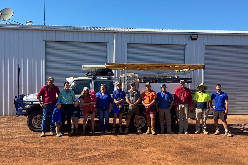 A mix of 14 men, women, and children stand outside in front of a shed and ute. 