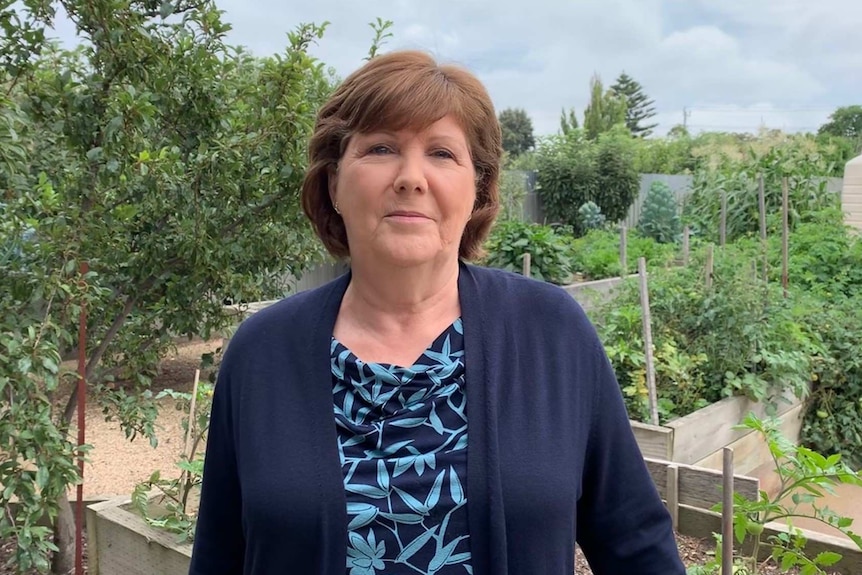 A woman with short brown hair, wearing a navy blue cardigan and light blue top stands in a lush green vegetable garden.
