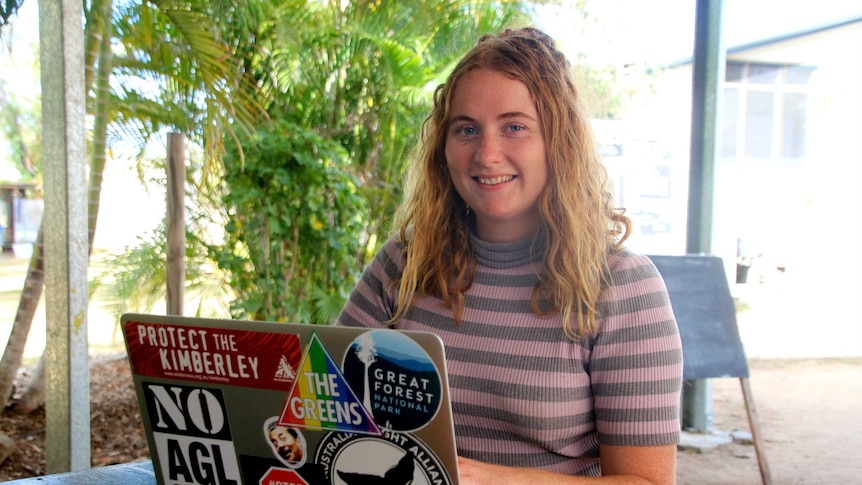 A girl with a striped jumper sits at a table with a laptop with stickers on it