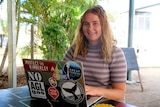 A girl with a striped jumper sits at a table with a laptop with stickers on it