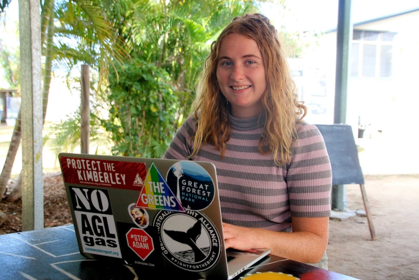 A girl with a striped jumper sits at a table with a laptop with stickers on it