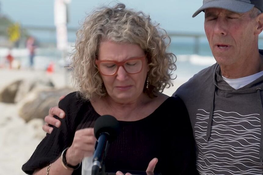 An emotional woman and man speak into a microphone at a beach setting.